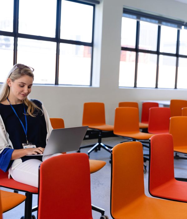 Front view of a Caucasian woman with long blond hair, wearing smart clothes, sitting on a chair in an empty modern meeting room, using a laptop.