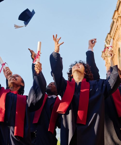 Group of students in gowns tossing their hats up while standing outdoors near the university