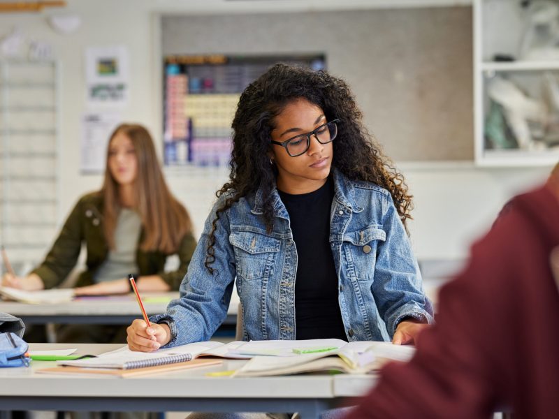African american girl reading and taking notes during a lesson in classroom. Multiethnic young student with eyeglasses studying and doing test in classroom at high school. Black female teen in casual during exam in secondary school.