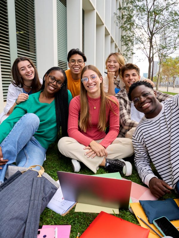 Portrait of diverse young group student friends posing happy looking smiling at camera outdoors. Cheerful Generation z classmates sitting together with laptop and workbooks in university campus park