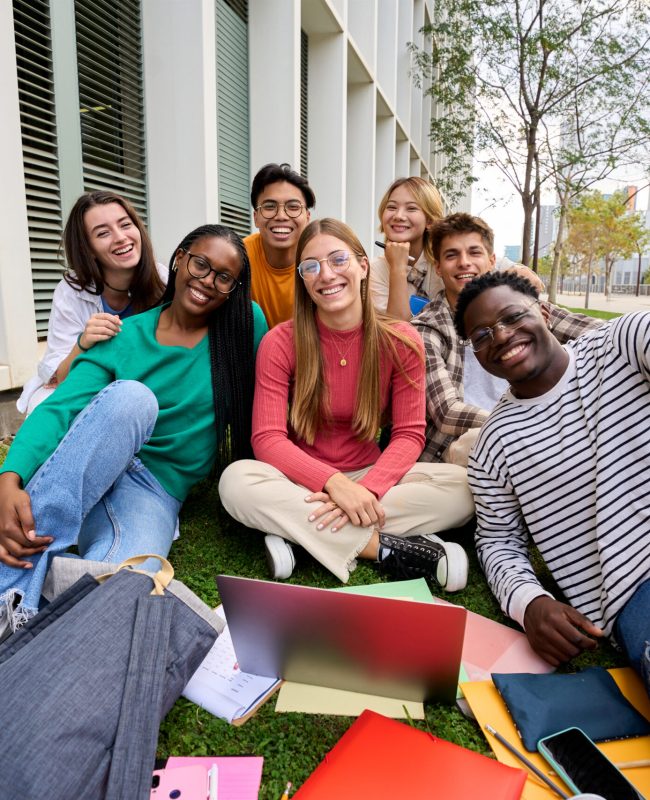 Portrait of diverse young group student friends posing happy looking smiling at camera outdoors. Cheerful Generation z classmates sitting together with laptop and workbooks in university campus park