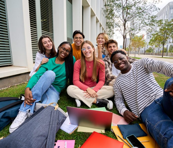 Portrait of diverse young group student friends posing happy looking smiling at camera outdoors. Cheerful Generation z classmates sitting together with laptop and workbooks in university campus park